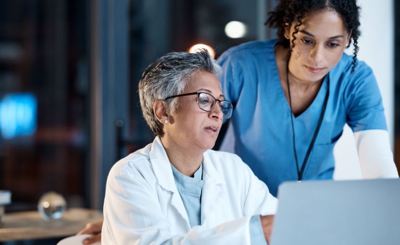 Two medical professionals - one wearing blue scrubs, the other wearing a white lab coat - look at a laptop screen together