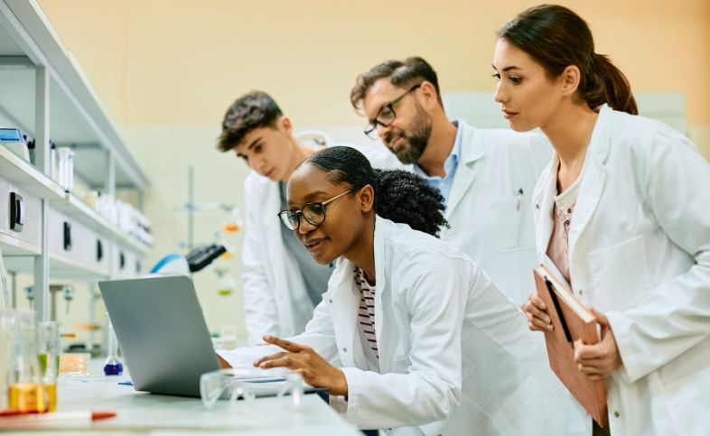 Group of scientists in white lab coats look around a laptop