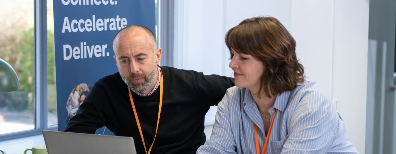 Deb and Ben talking together in the office. Connect, Accelerate, Deliver in the background