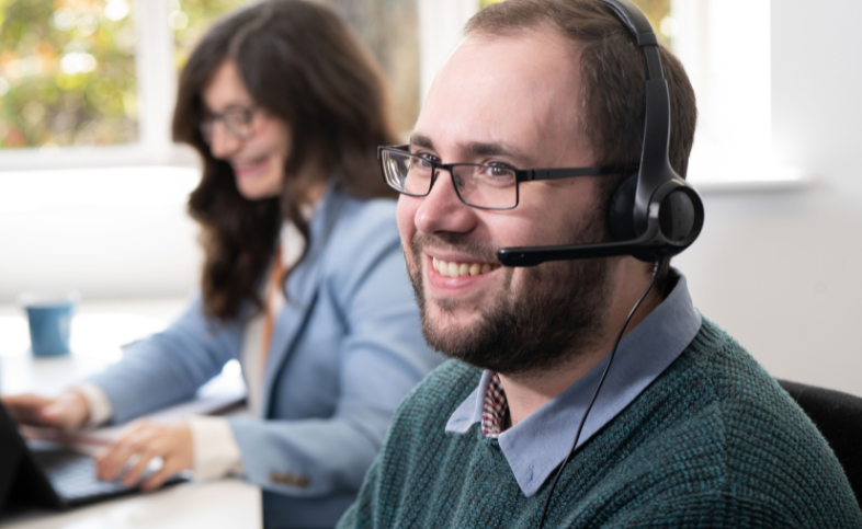 Luke smiles with headset on, Kristina works in the background