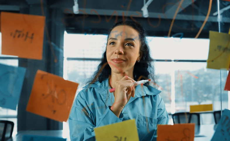 Lady stands in front of glass screen holding a pen. The screen is full of writing and post it notes with comments.