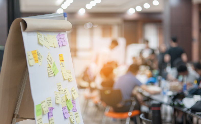 Close up shot of a flipchart with post it notes on, in the background a group sits round a table discussing ideas