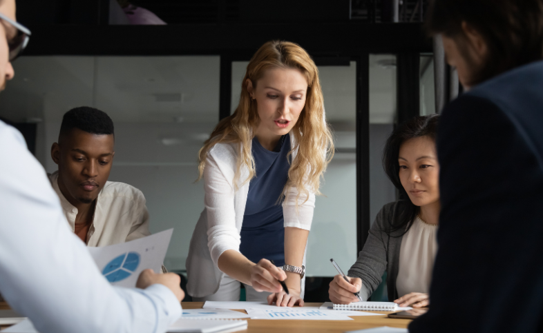 Formal team meeting with five individuals sat round a table looking at a piece of paper. The lady in the centre is leaning over the paper with a pen in her hand pointing
