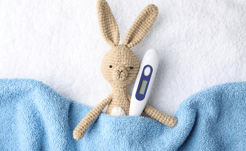 Toy knitted bunny is tucked under a blue towel with a thermometer next to it