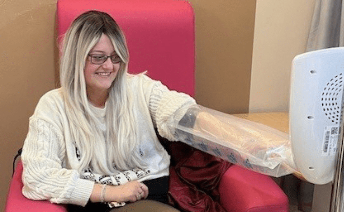 Lady sits in a big pink hospital chair, with her arm in a plastic bag as part of the airglove device