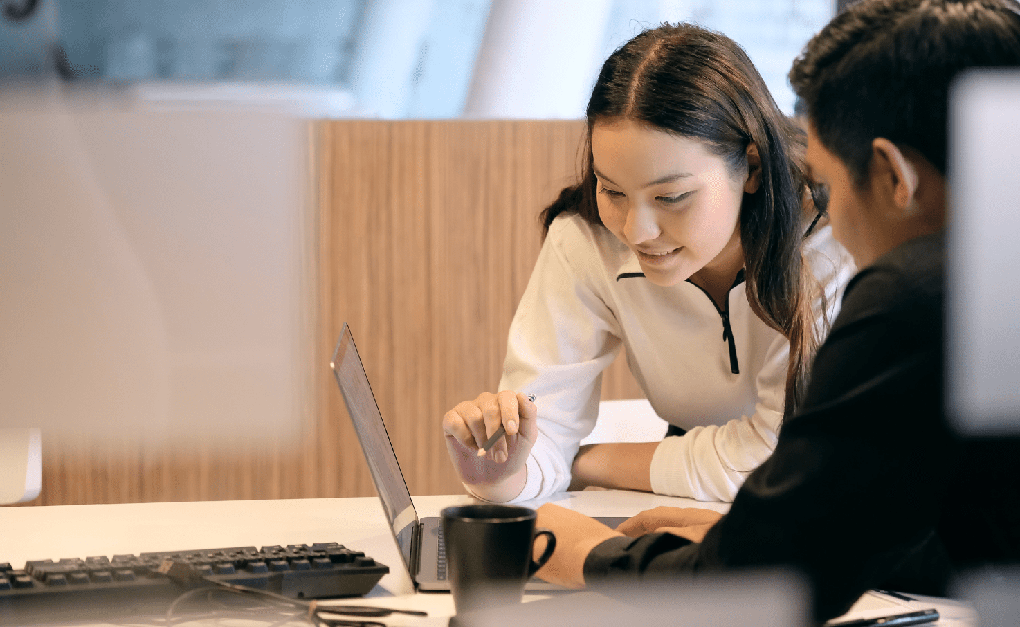 Two people work together in an office, one leans over to look at the other persons laptop