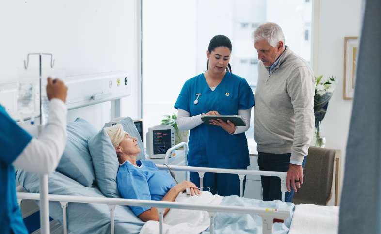 Female patient lays in hospital bed, husband talks to doctor at the side of the bed