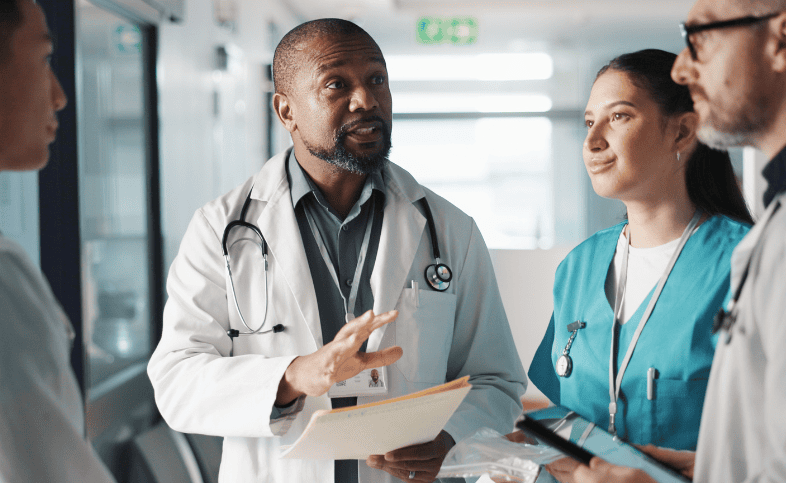 Hospital staff, one in a white lab coat and stethoscope and one in a pair of green scrubs talk, to other people join the conversation just out of the picture